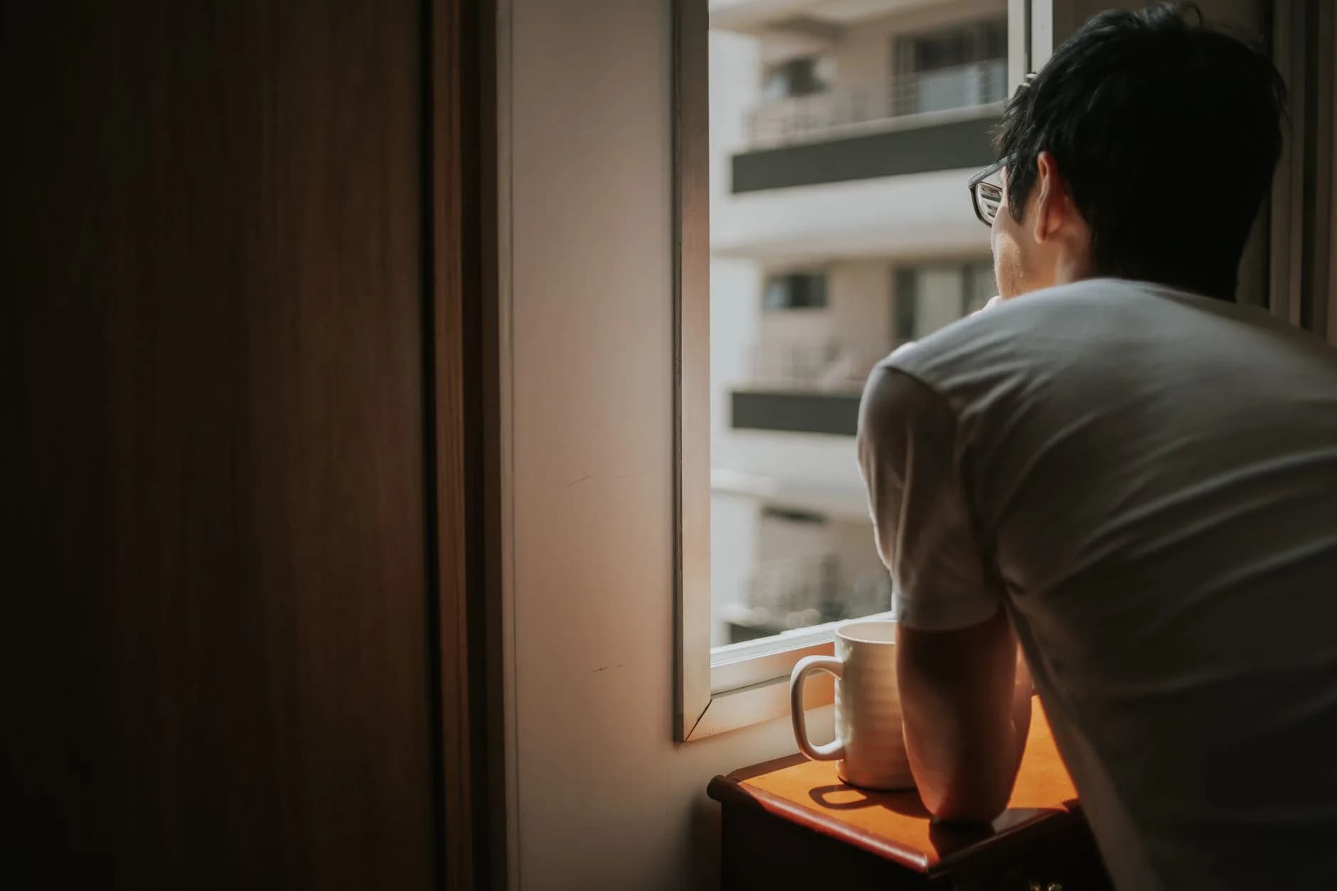 A man thoughtfully looks out a window with city buildings in view, holding a mug. · Фото: Letícia Alvares / Pexels