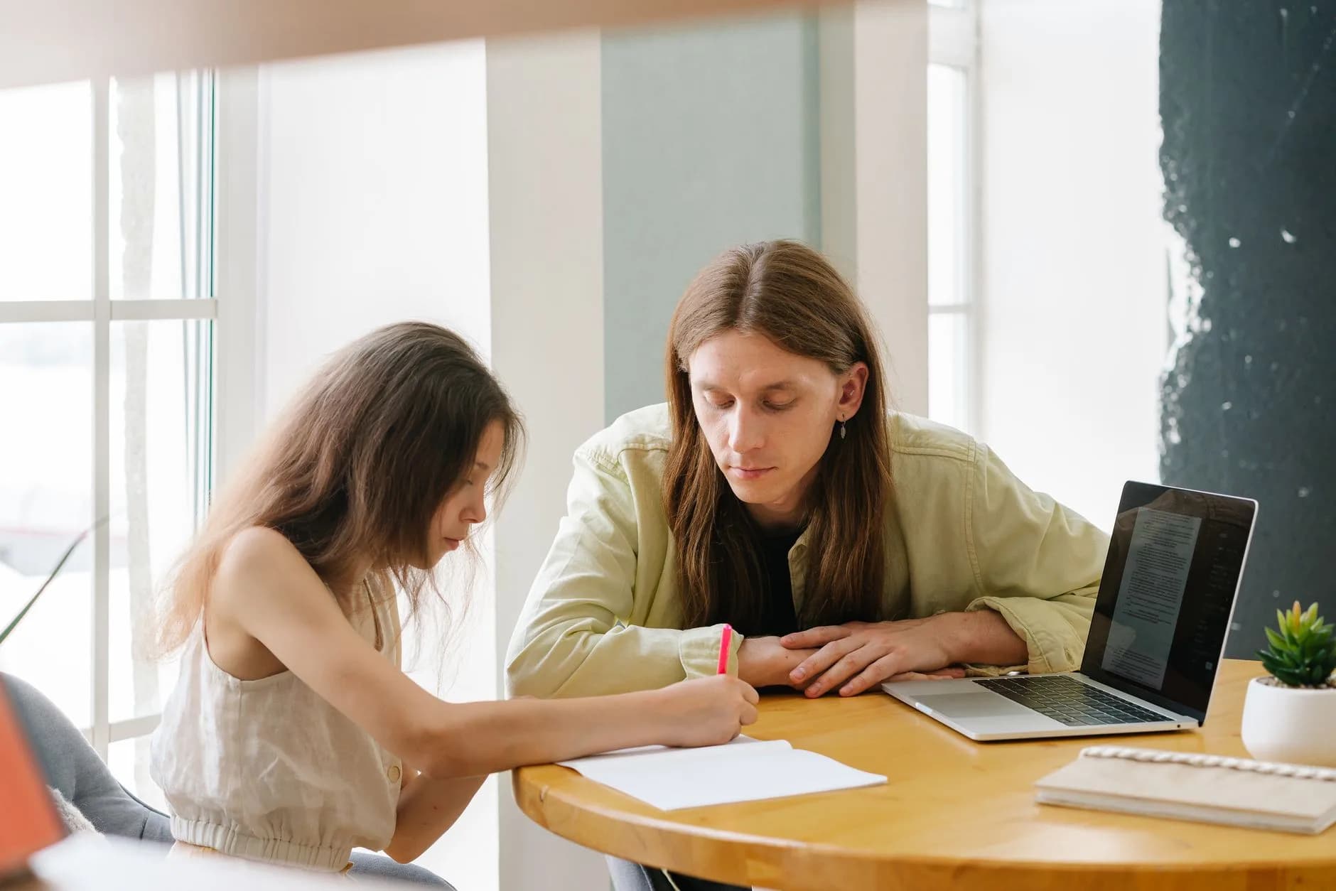 A father helps his daughter with homeschooling indoors, featuring a laptop and study materials. · Фото: Mikhail Nilov / Pexels